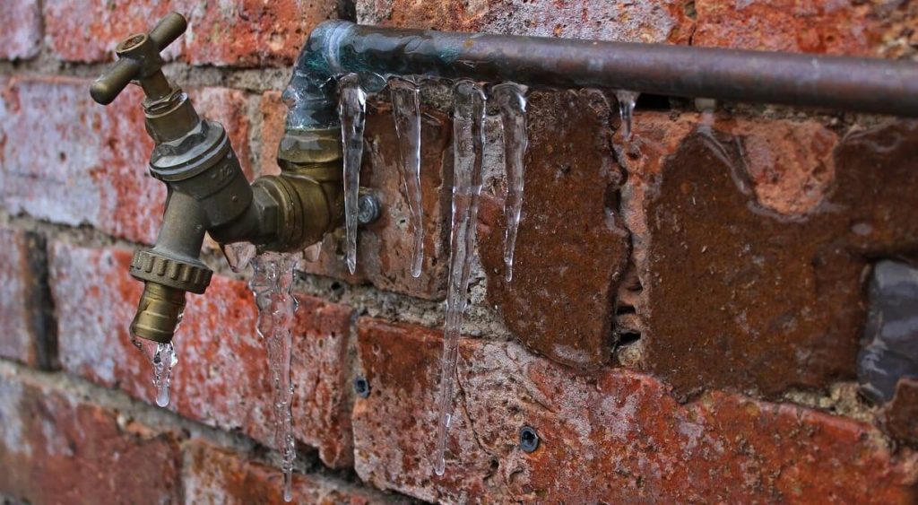 Red brick wall with a metal frozen pipe and tap attached with icicles hanging off it